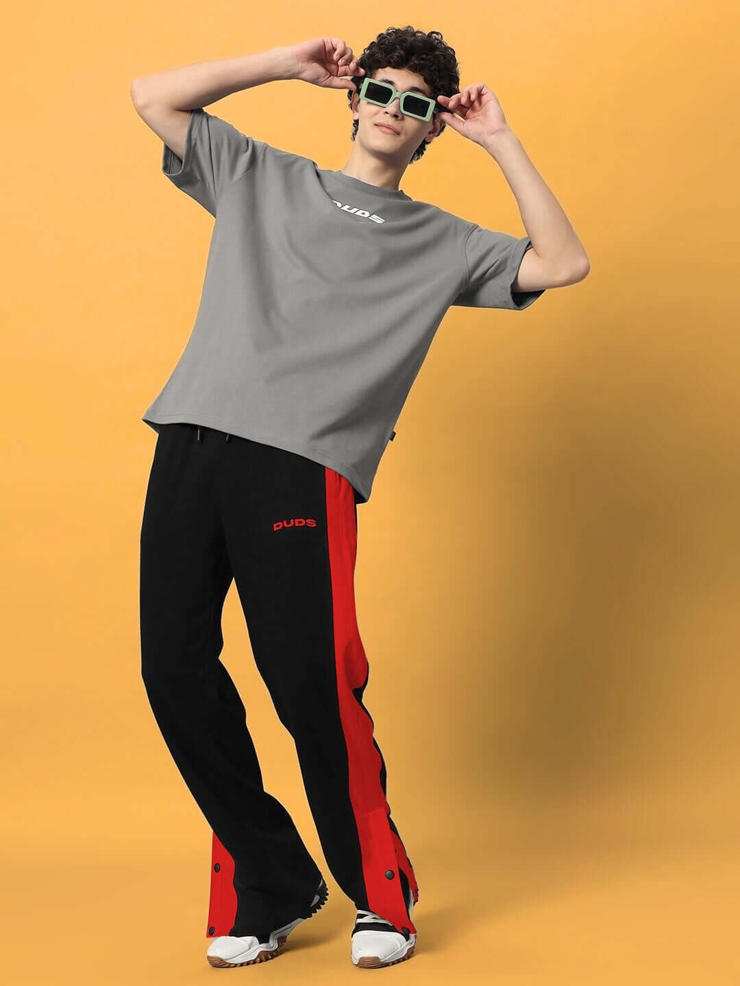 Model wearing black-red Flurry Contrast Joggers by DUDS, styled with a grey t-shirt and sunglasses, standing against an orange background.