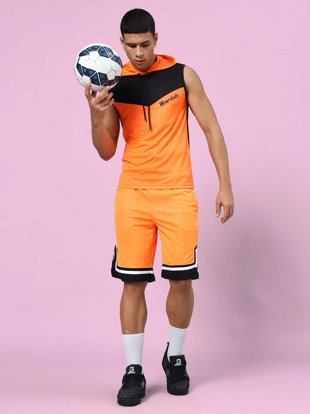 Man in FRANKY GYM CO-ORD SET ORANGE BLACK holding a soccer ball, wearing an athletic sleeveless hoodie and shorts, stylish sportswear.