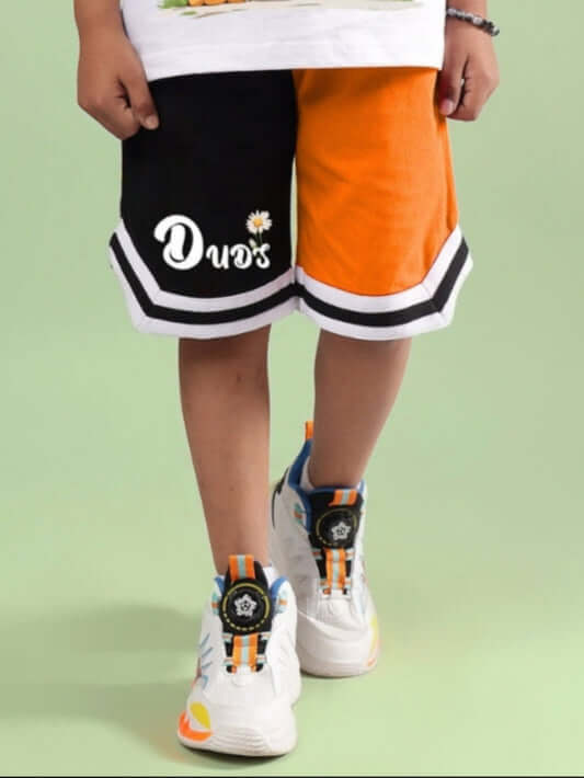 FRIENDSHIP BUCKET SHORTS FOR BOYS & GIRLS BLACK-ORANGE worn by a child, white shirt, colorful sneakers, green background, Duds logo visible.