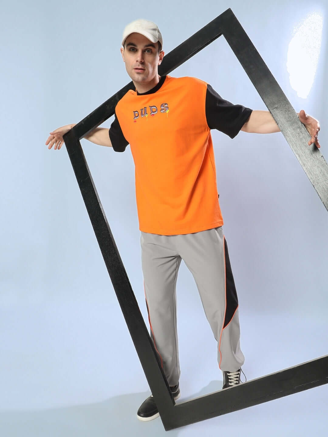 Model wearing grey relaxed fit cargo pants and orange DUDS shirt, posed creatively with a black frame.