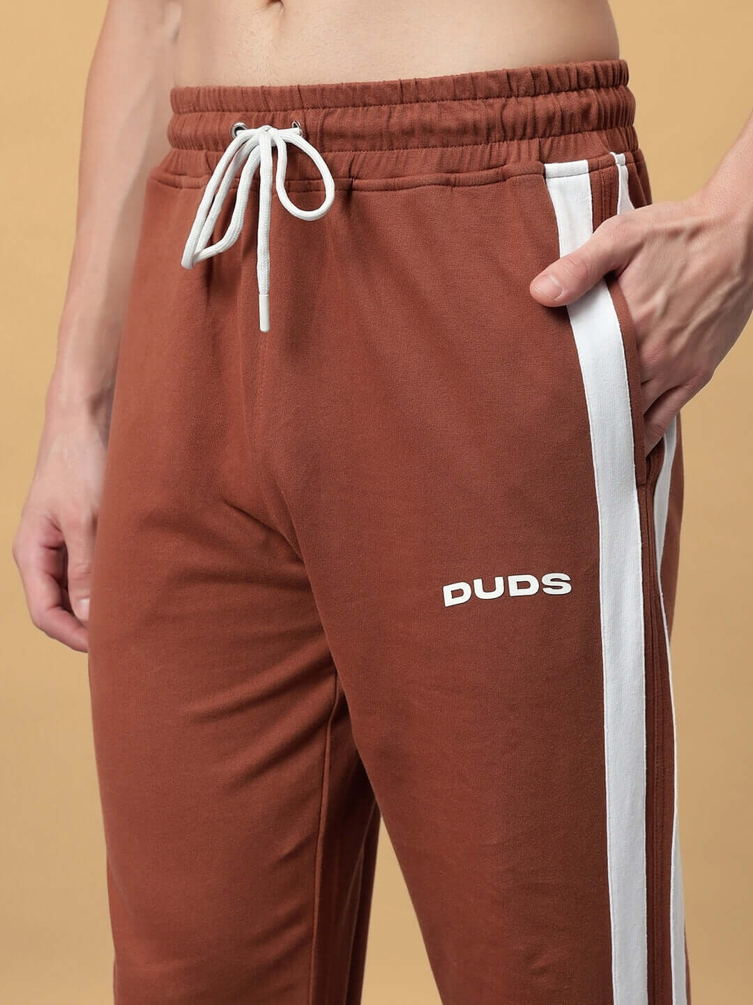 Close-up of brown FUTURE ICON 2 STRIPE JOGGER with white stripes and DUDS logo, highlighting elastic waistband and drawstring.