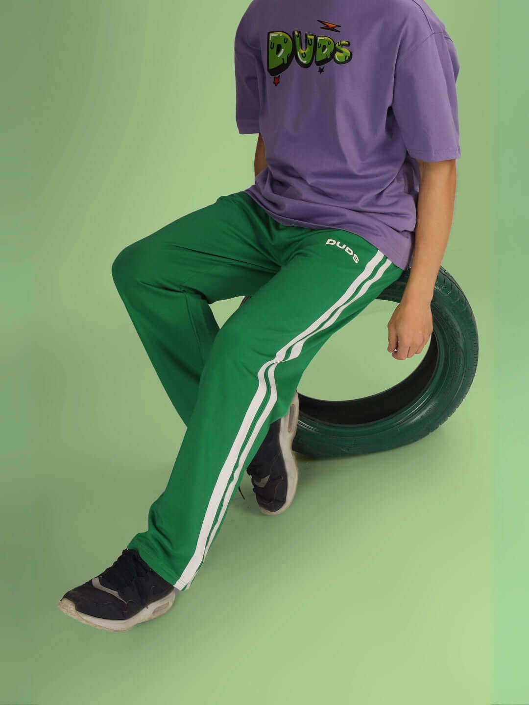 Model wearing Future Icon 2 Stripe Jogger in green, paired with a purple DUDS t-shirt, sitting on a tire. Custom fit available.
