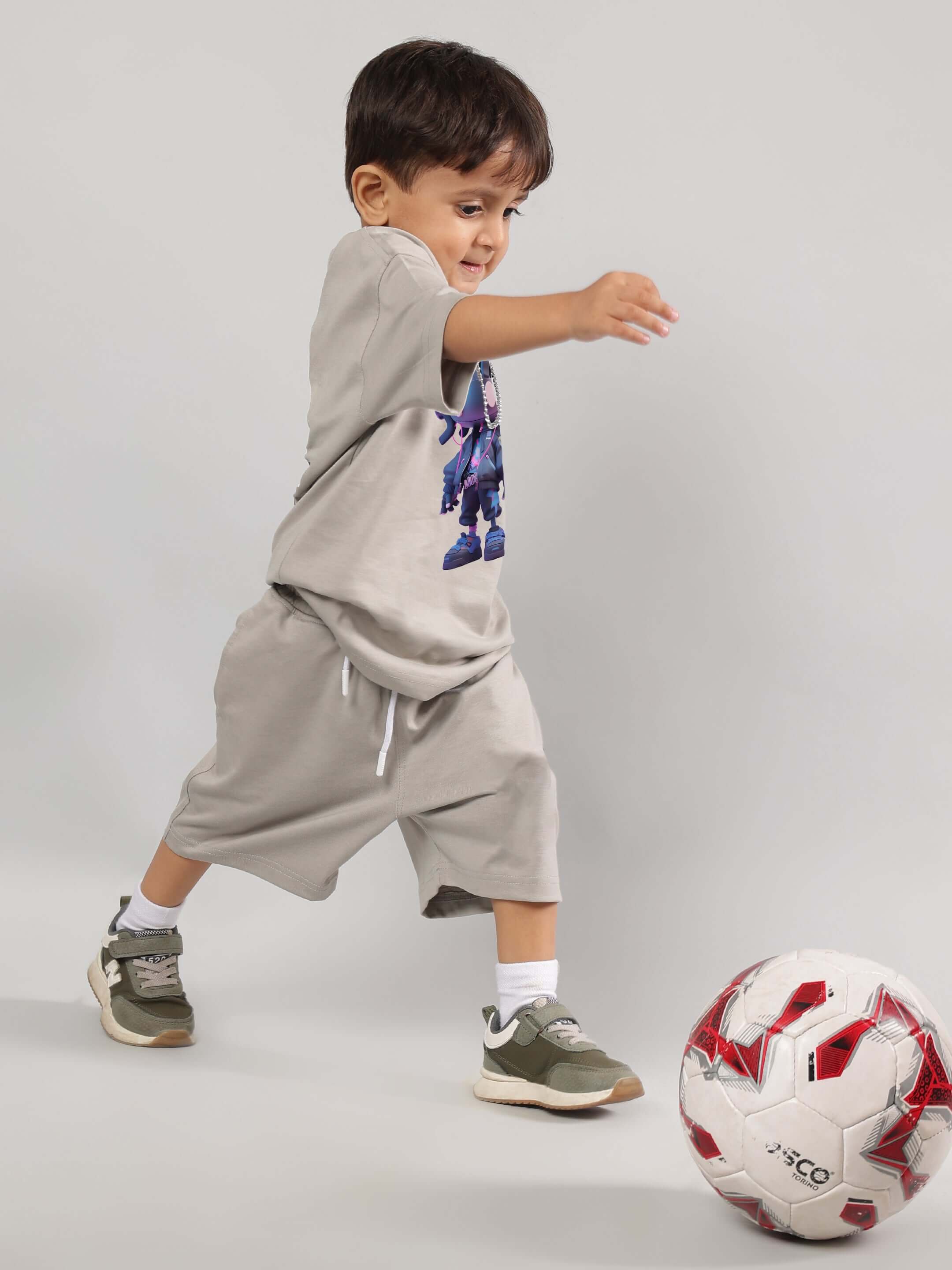 Young boy wearing a grey futuristic kids t-shirt with tech-inspired graphic, kicking a soccer ball indoors.