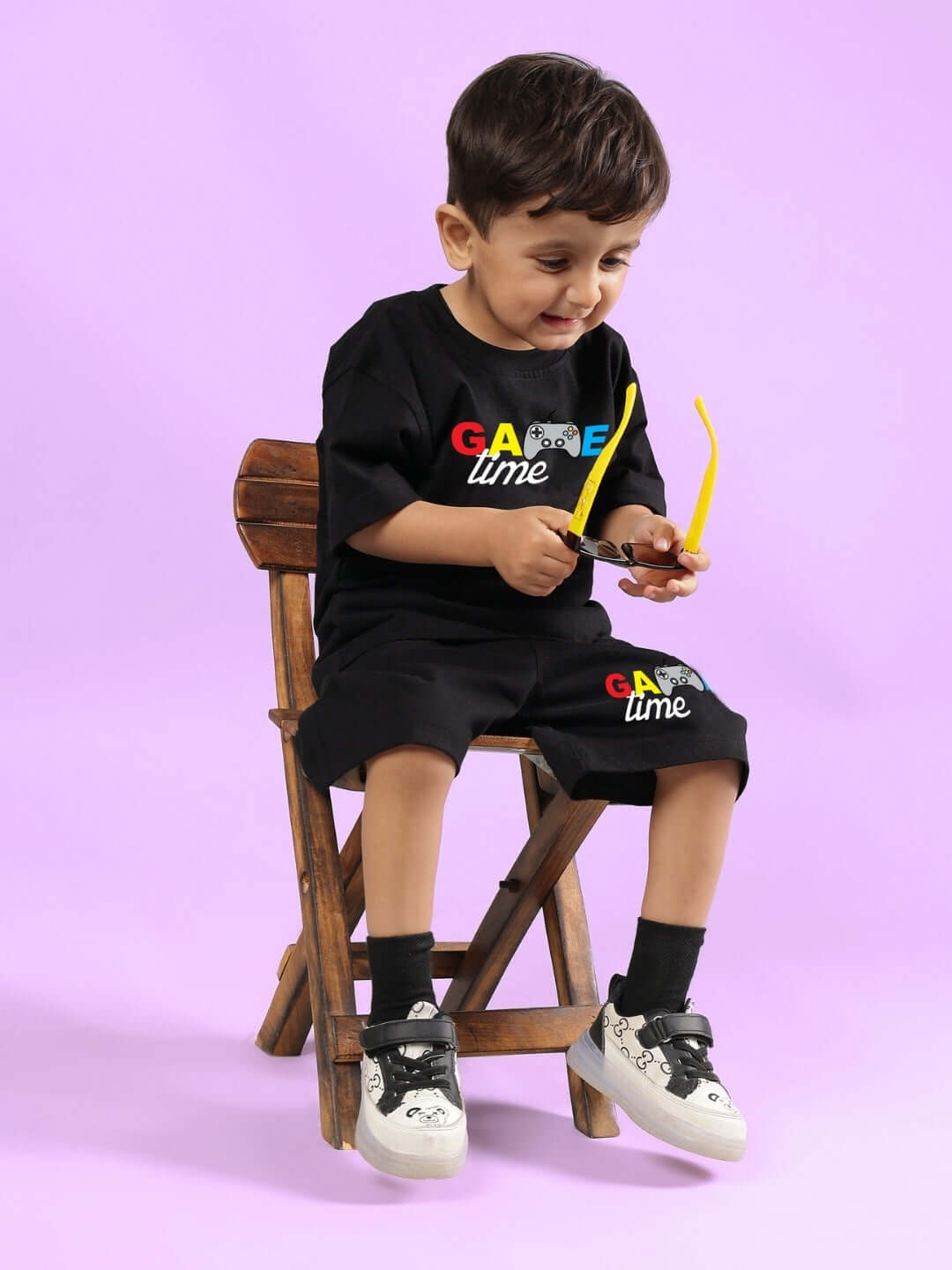 Child wearing GAME FINISHER SHORTS FOR BOYS & GIRLS BLACK, sitting on a wooden chair holding sunglasses.
