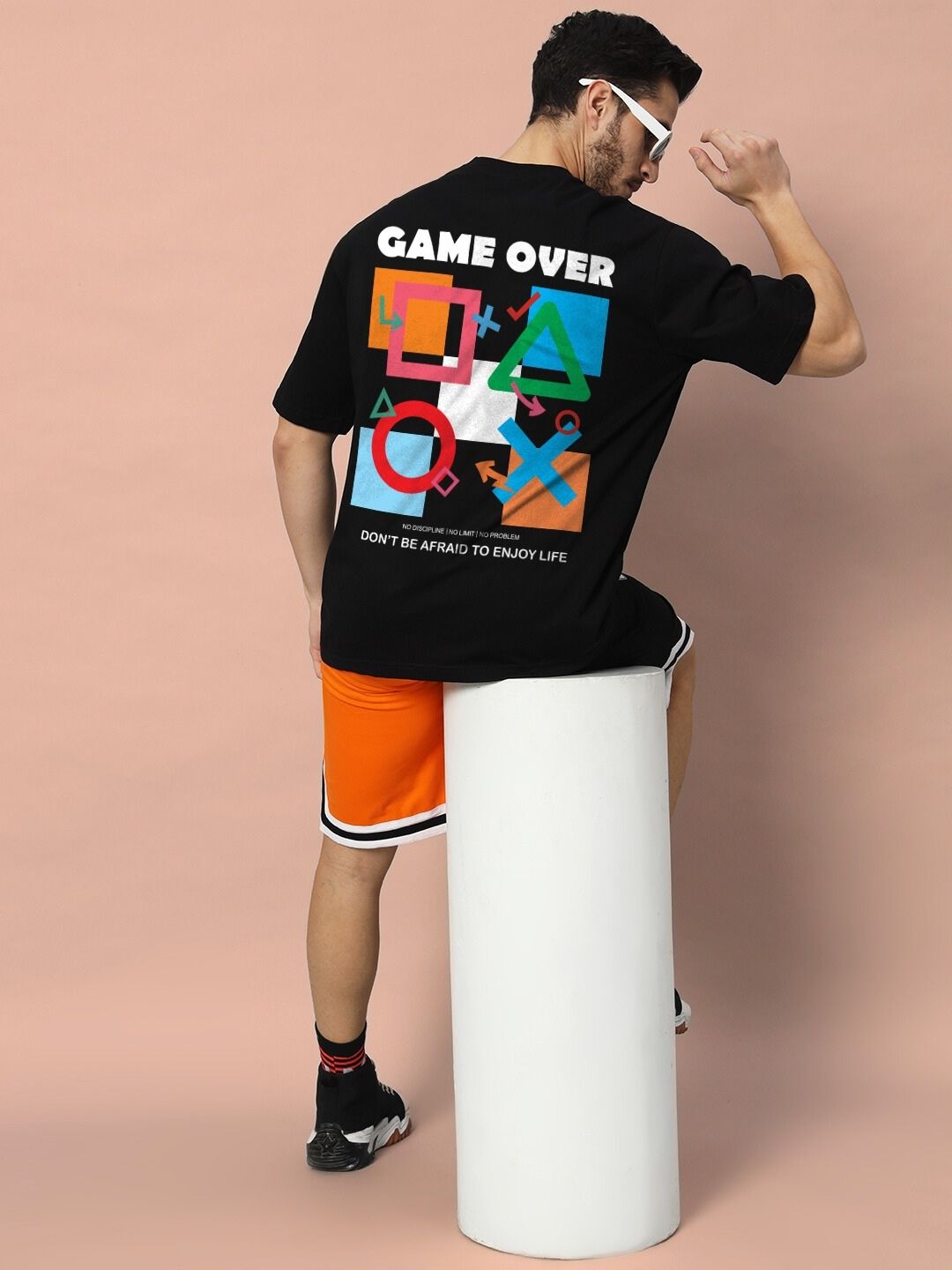 Model wearing GAME-OVER OVER-SIZED T-SHIRT BLACK with graphic print, sitting on a white stool.