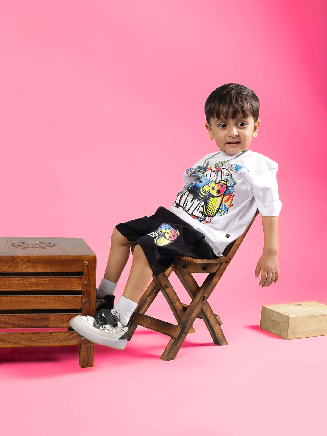 Child wearing GAME TIME CO-ORD FOR BOYS & GIRLS WHITE BLACK clothing, sitting on chair in colorful outfit, pink background.