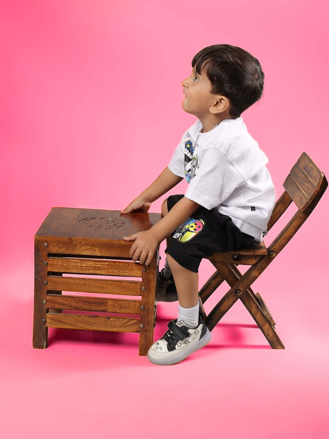 Boy in GAME TIME CO-ORD FOR BOYS & GIRLS WHITE BLACK outfit, sitting on a wooden chair with pink backdrop, looking upward.