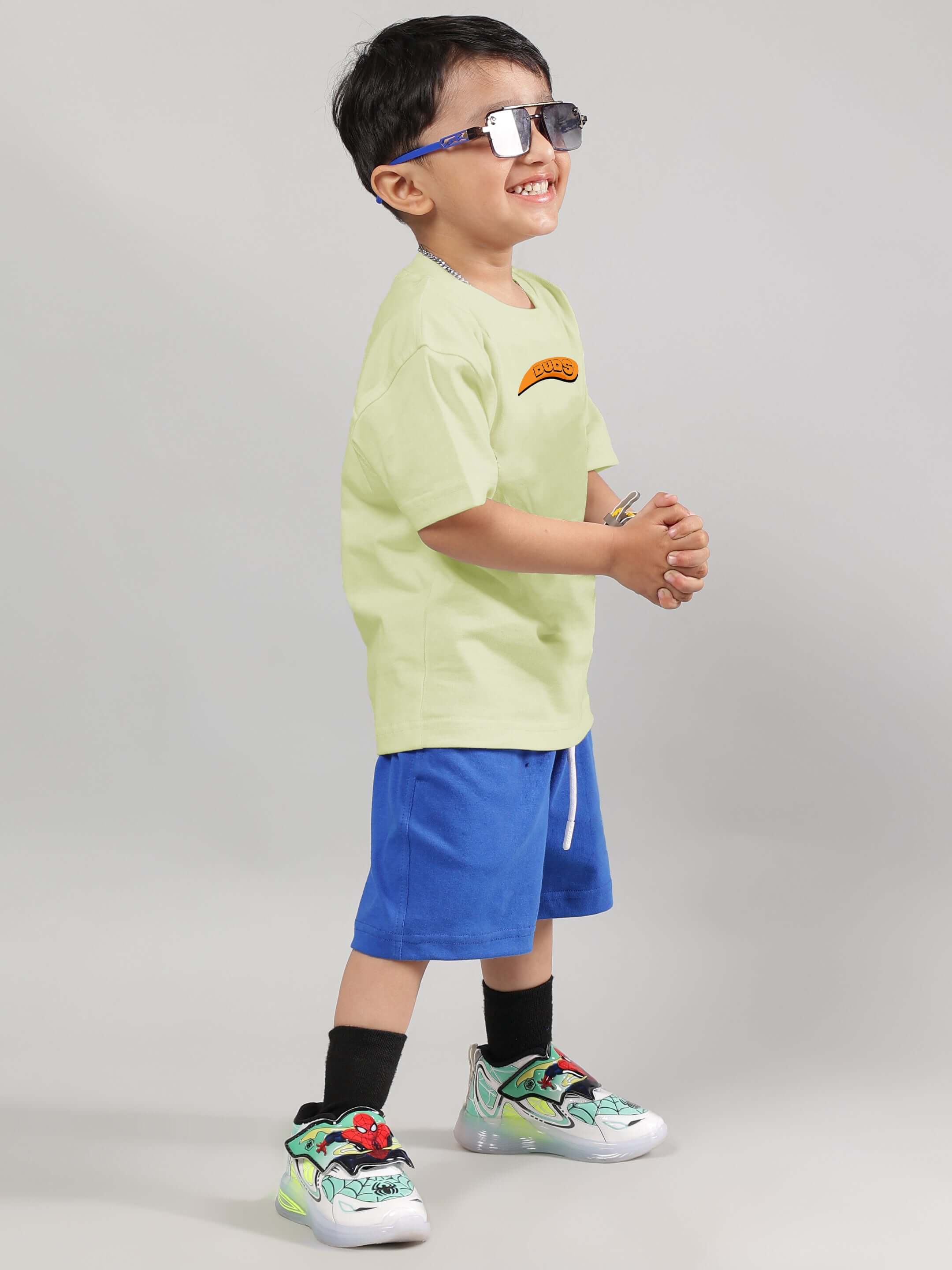 Young boy wearing Garfield co-ord set with celery green T-shirt and blue shorts, smiling and wearing stylish sneakers.