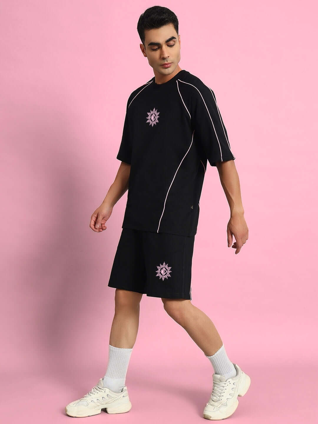 Model wearing GLARE REGULAR FIT SHORTS BLACK paired with matching black t-shirt and white sneakers on pink background.