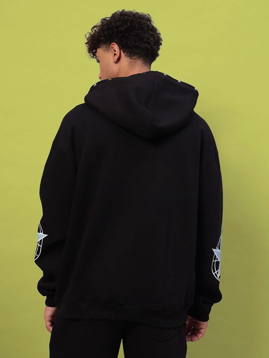 Back view of a person wearing the GLOBAL COTTON FLEECE HOODIE BLACK, showing detailed design on sleeves, against a green background.