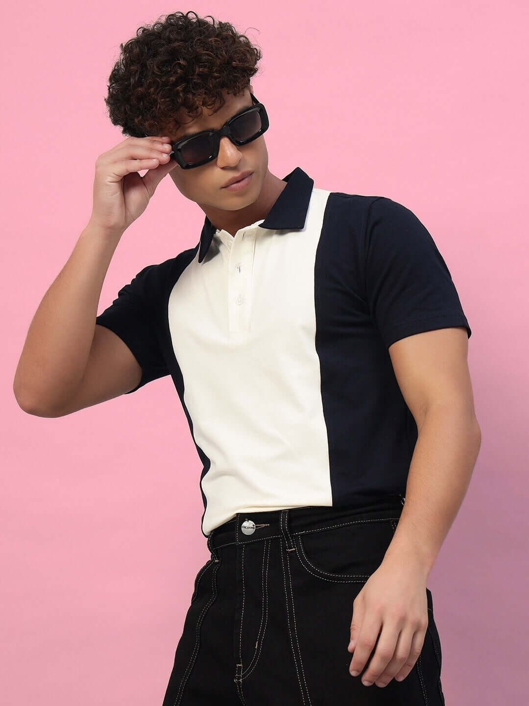 Model wearing a black and white collared t-shirt with dark jeans against a pink background, adjusting sunglasses.