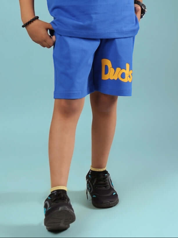 Child wearing GO MOTION SHORTS FOR BOYS & GIRLS ROYAL BLUE with sneakers on a blue background.