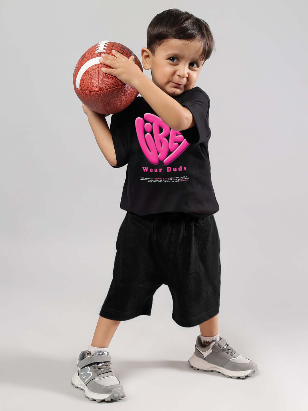 Young child wearing a black 'Good Vibes' t-shirt, holding a football, showcasing DUDS sportswear for boys and girls.
