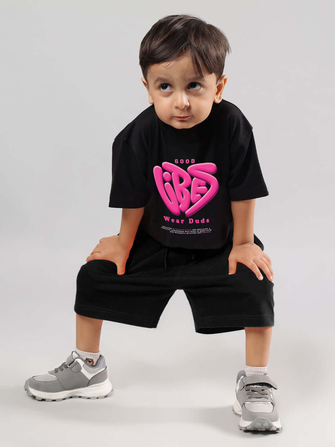 Toddler wearing a black 'Good Vibes' t-shirt from DUDS with pink lettering, paired with shorts and sneakers, sitting on a stool.