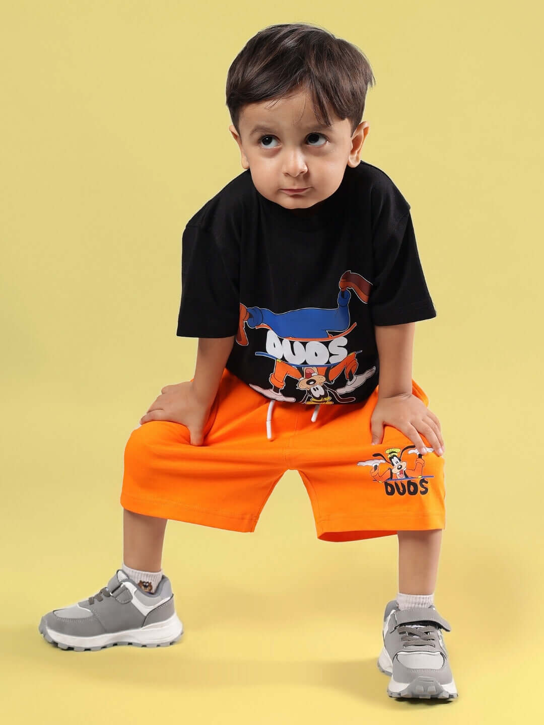 Child wearing GOOFY SUMMER CO-ORD FOR BOYS & GIRLS BLACK-ORANGE, featuring black t-shirt and orange shorts, sitting on yellow background.