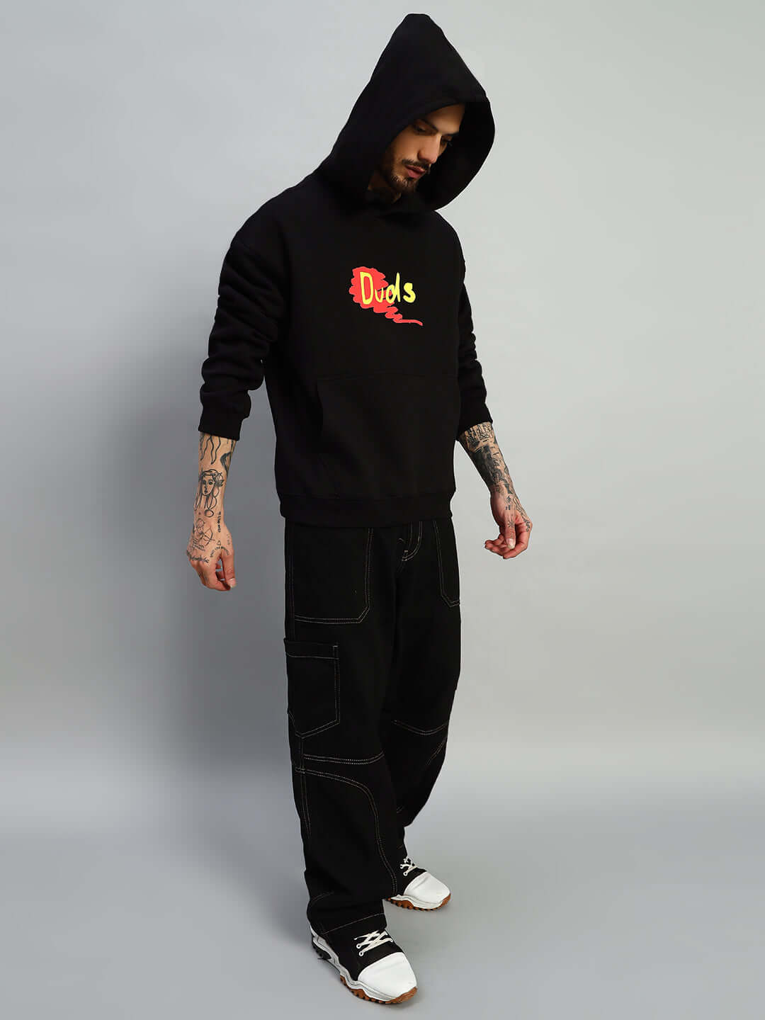 Person wearing black oversized hoodie with graphic design, paired with black pants and white shoes, showcasing street fashion style.