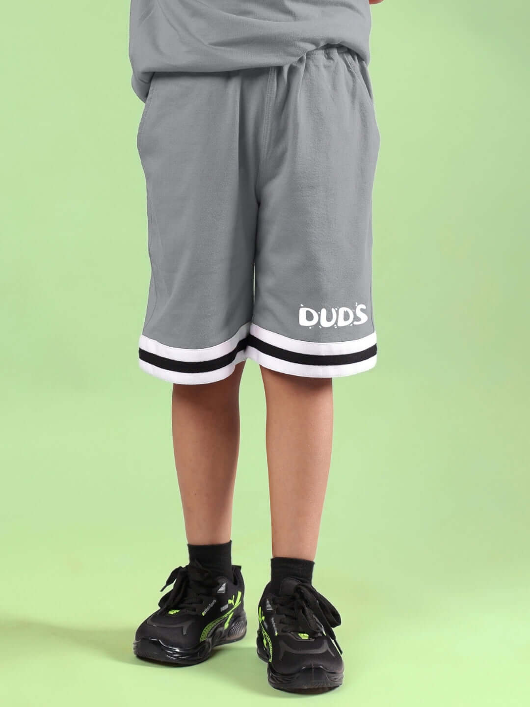 Child wearing grey LETS GO PLAY printed shorts for boys and girls, featuring DUDS logo, against a green background.