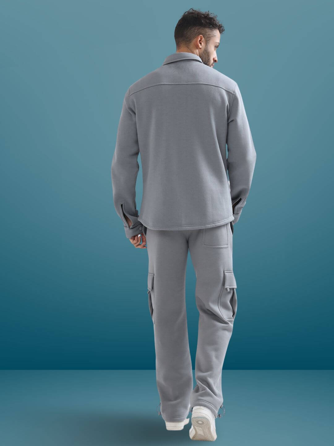 Back view of a model wearing grey relaxed fit cargo pants with matching jacket on a blue background.