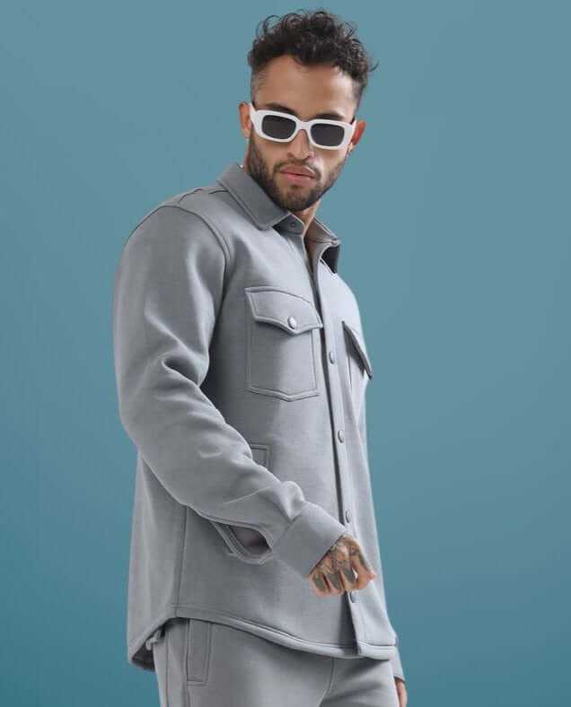 Man wearing a grey relaxed fit overshirt, sunglasses, and casual trousers against a blue background.