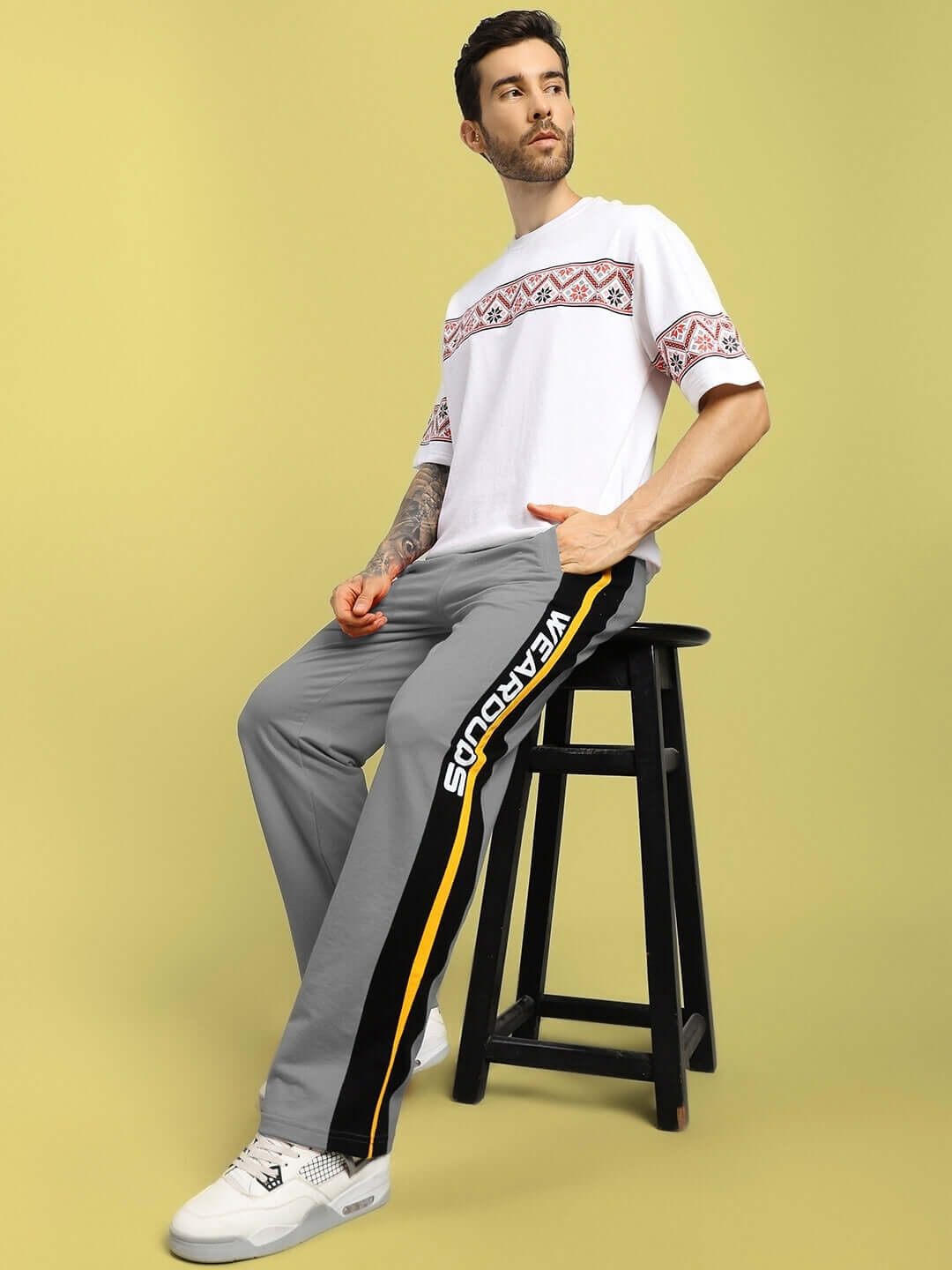 Model wearing Roger relaxed-fit grey joggers with vibrant side stripe, sitting on stool in casual style.
