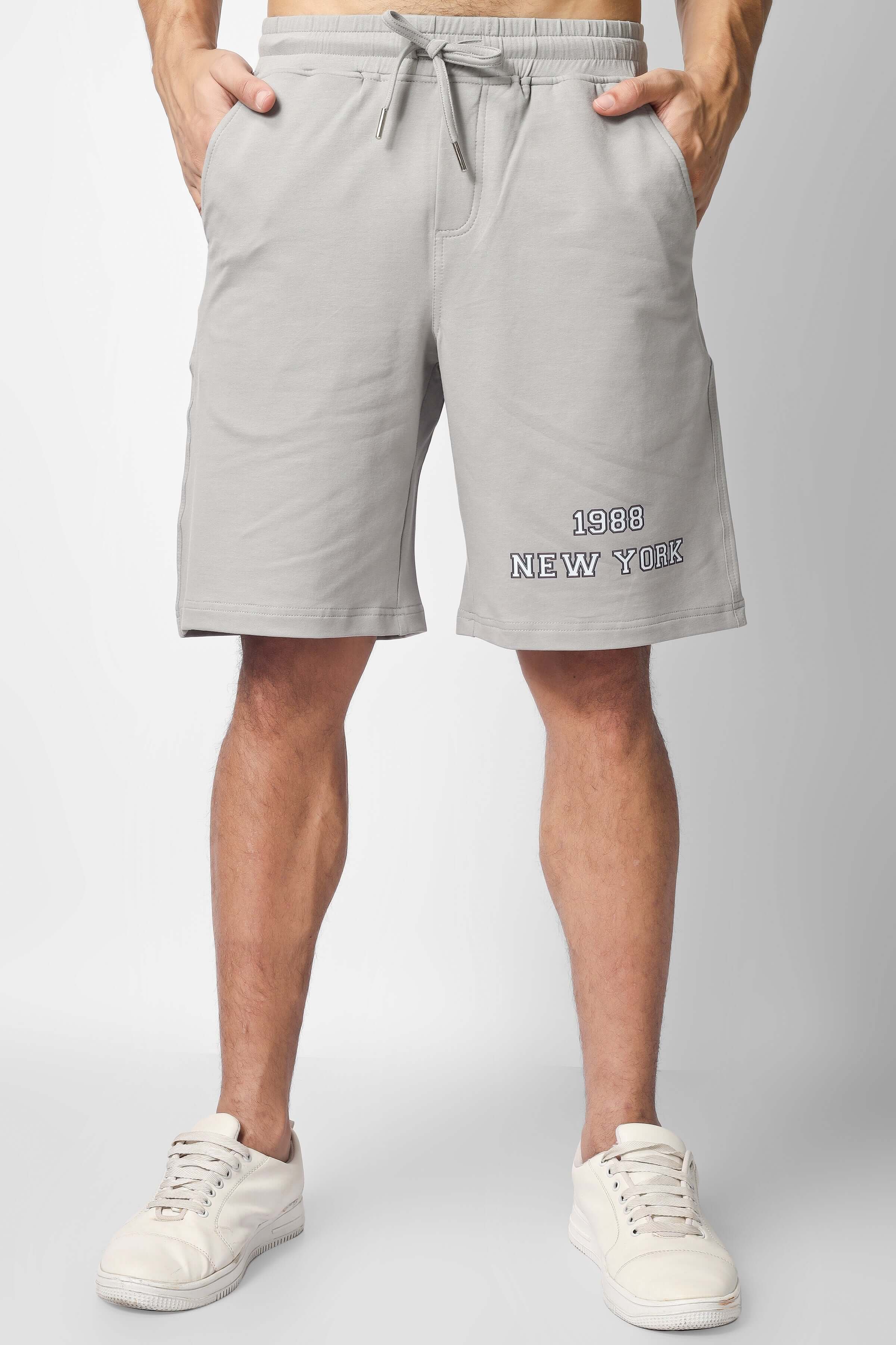 Man wearing grey shorts with '1983 New York' embroidery, white sneakers; part of the Brooklyn Co-Ord Set in white and grey.