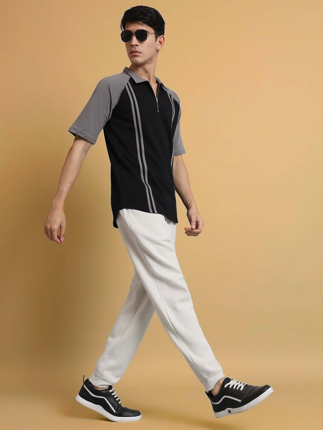 Man wearing black grey stripe raglan t-shirt with white pants, styled with sunglasses and sneakers, posing against a beige background.