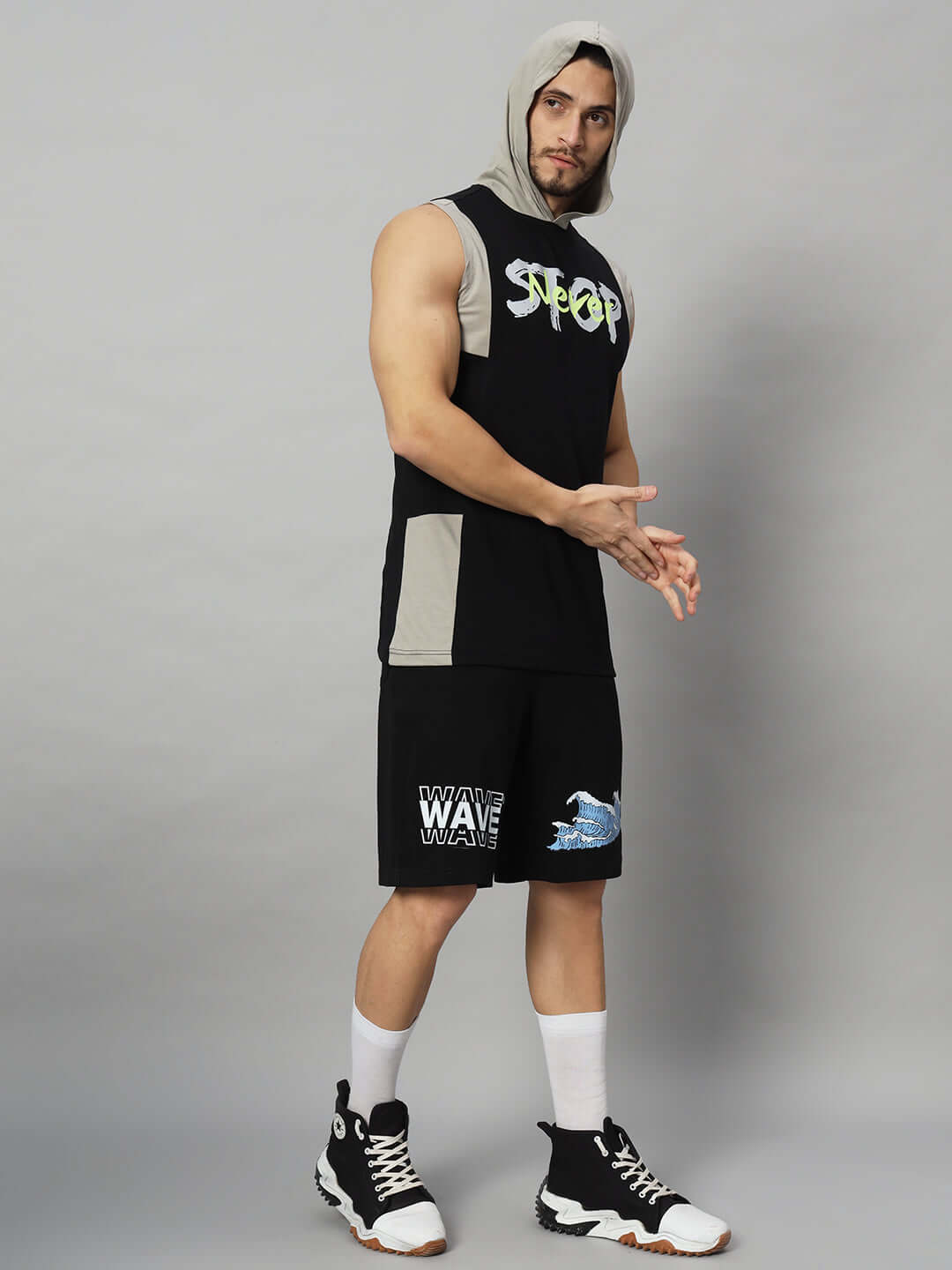 Man wearing stylish gym wear with hooded sleeveless top and shorts, showcasing a sporty look on a gray background.