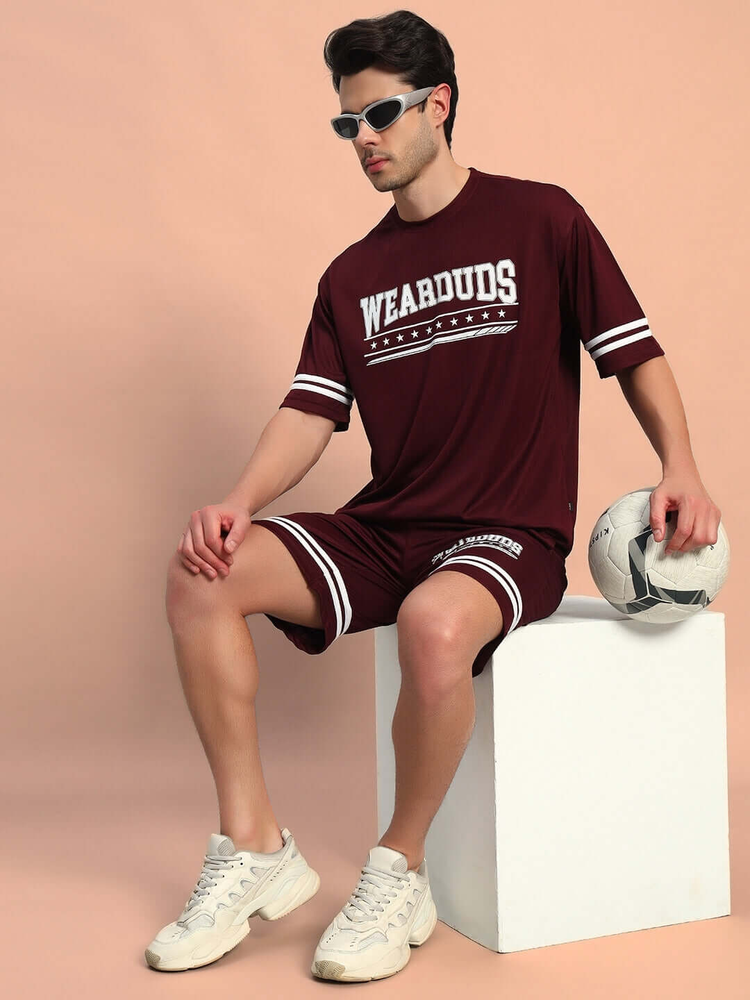 Man wearing GROOVY COLORBLOCK CO-ORD SET WINE by WEAR DUDS with retro sunglasses, sitting with a soccer ball, embodying vibrant streetwear style.