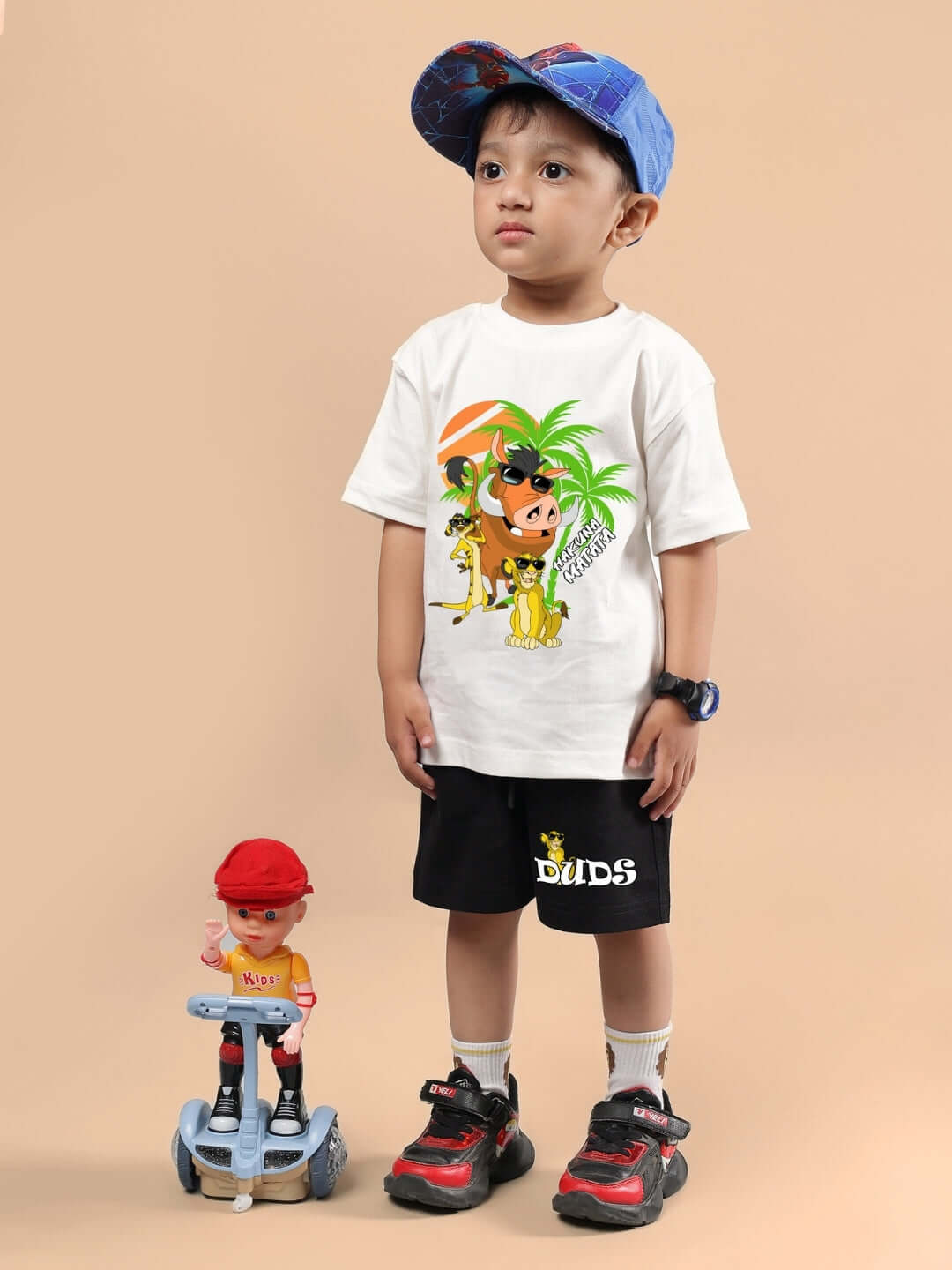 Child wearing HAKUNA MATATA SHORTS FOR BOYS & GIRLS BLACK with a white t-shirt, cap, and sneakers, standing next to a toy.