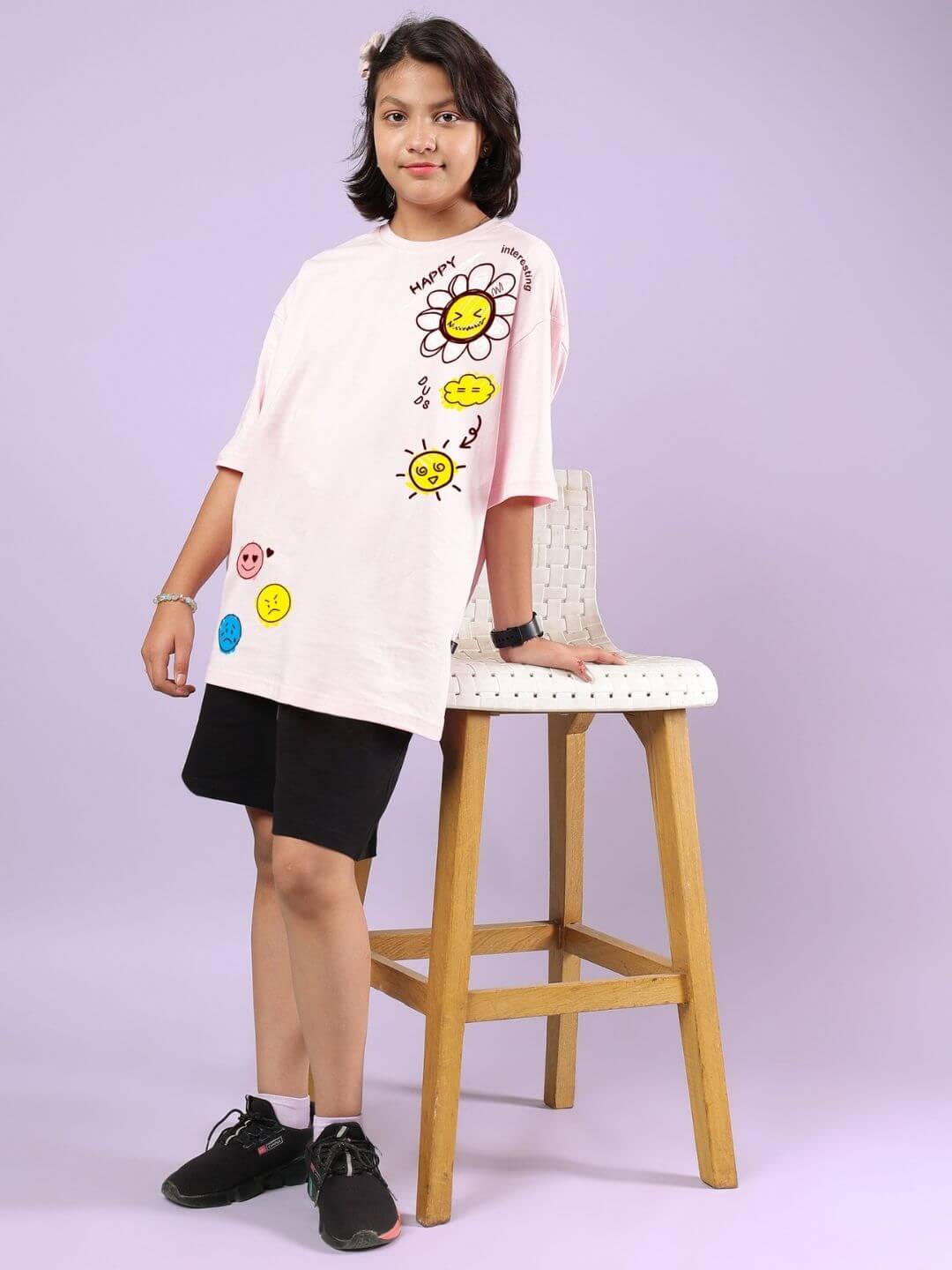 Child wearing Happy Interesting Co-ord Set in pink-black, featuring playful sun and flower designs, standing by a chair.