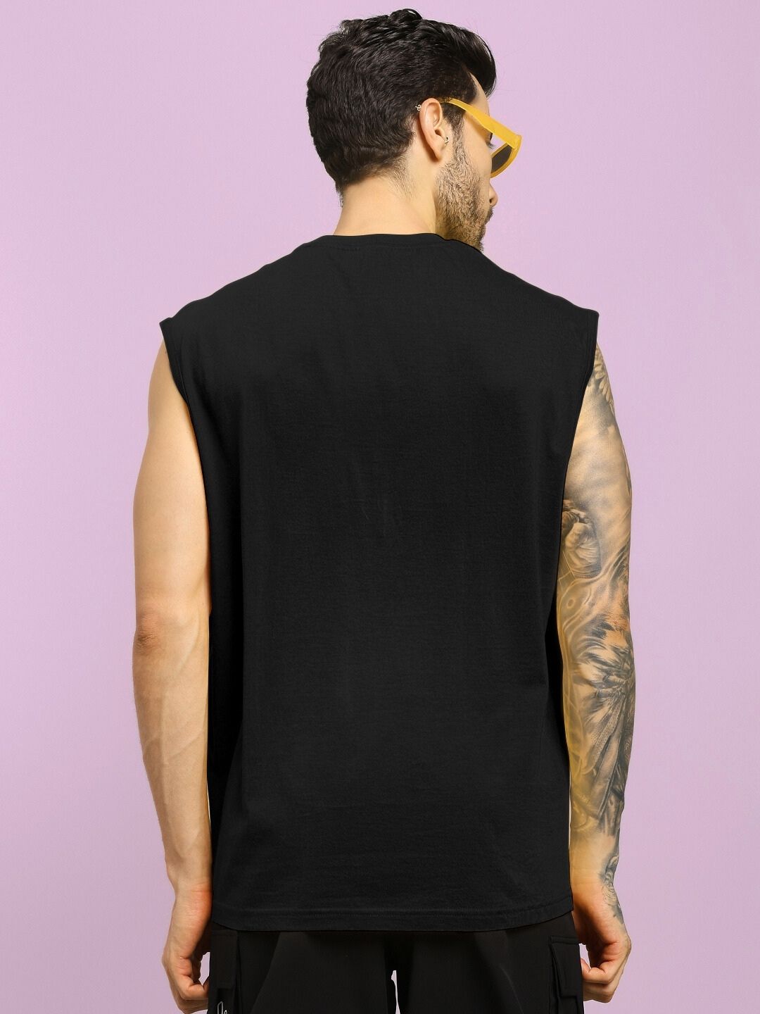 Rear view of a person wearing a HAPPY SKULL PRINTED GYM T-SHIRT BLACK, showcasing an arm tattoo and yellow sunglasses against a pink backdrop.