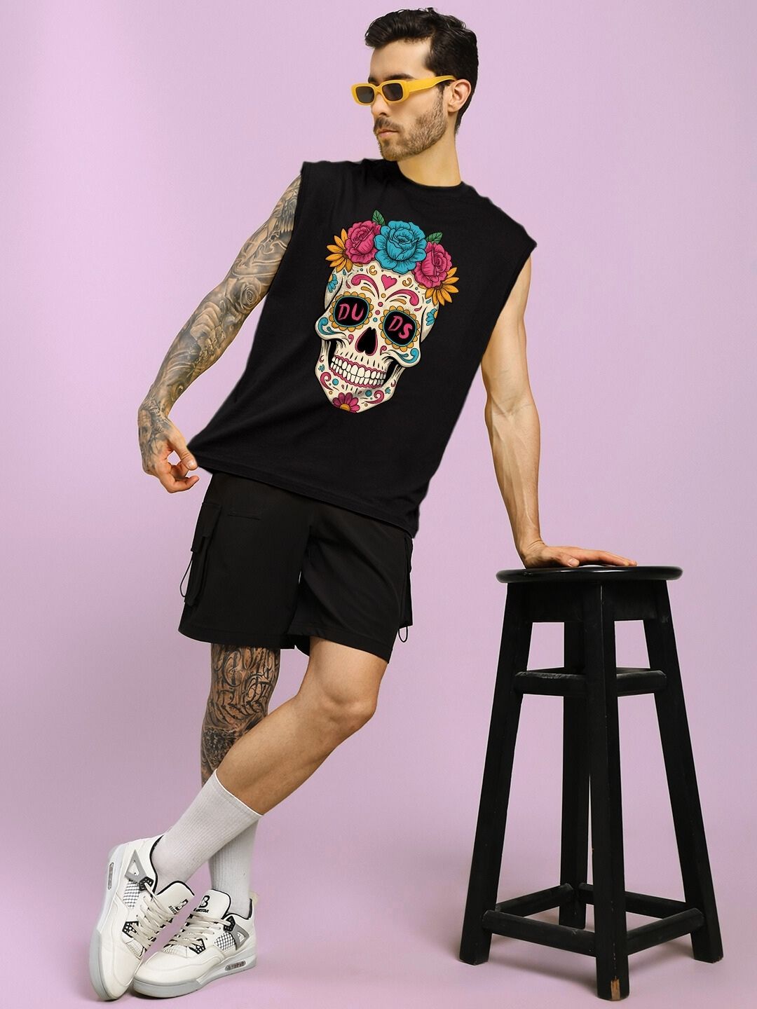 Man in a HAPPY SKULL PRINTED GYM T-SHIRT BLACK leaning on a stool, showcasing a colorful skull design, against a pink background.
