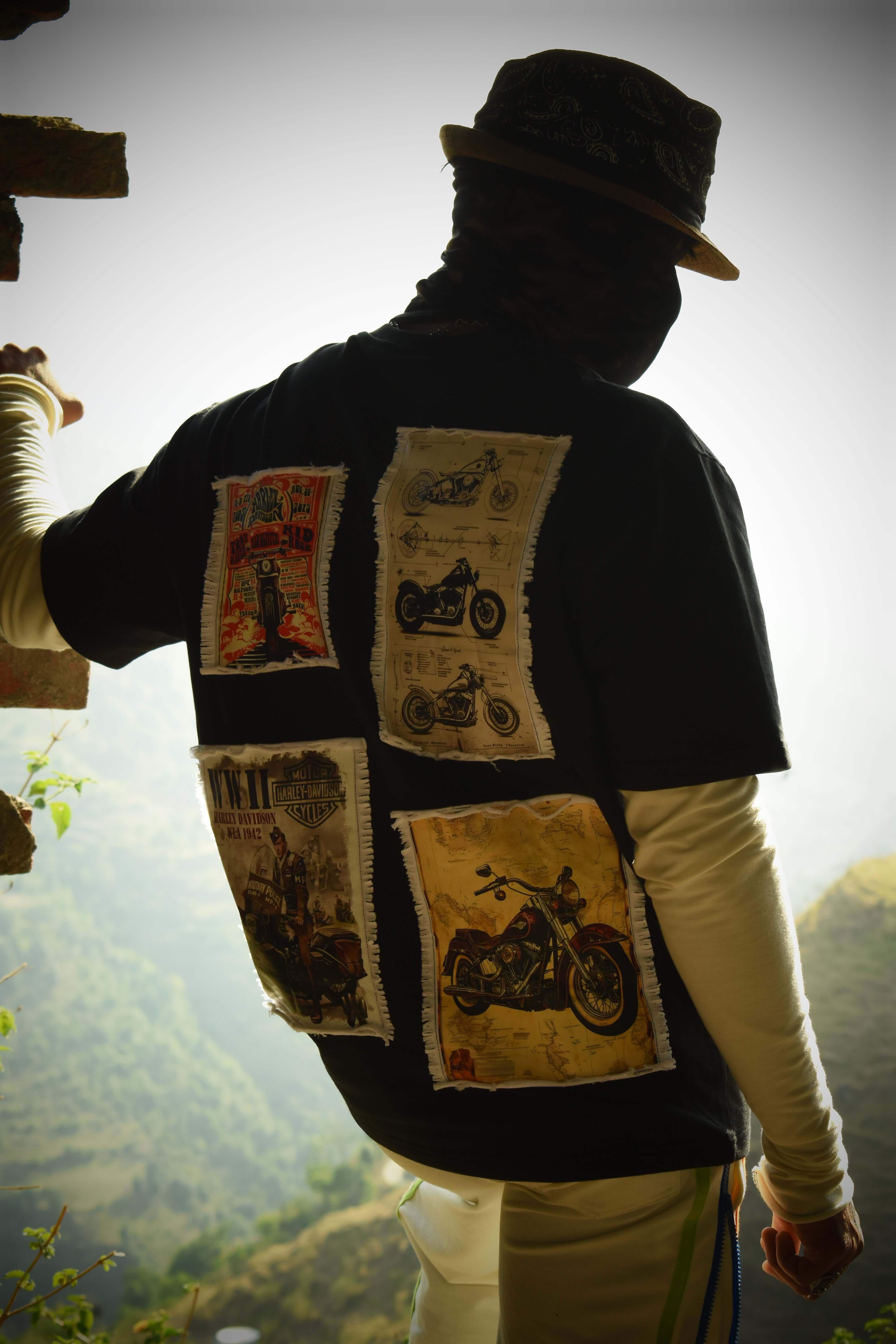 Person wearing HARLEY DESIGNER OVER-SIZED T-SHIRT BLACK with graphic motorcycle prints, standing outdoors with scenic view.