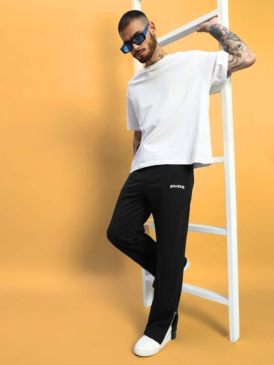Man wearing Hershey side slit joggers in black by DUDS; casual style with white t-shirt and sunglasses, leaning on a ladder.