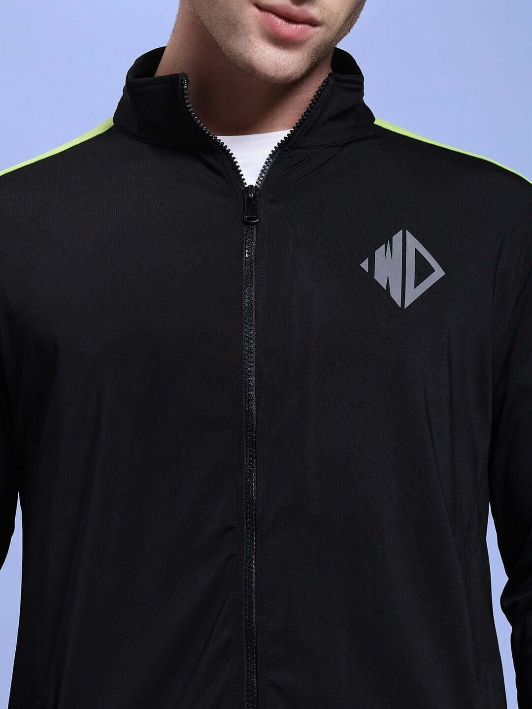 Close-up of a man wearing the HIGH PERFORMANCE JACKET IN BLACK with a zippered front and logo on the chest against a blue background.