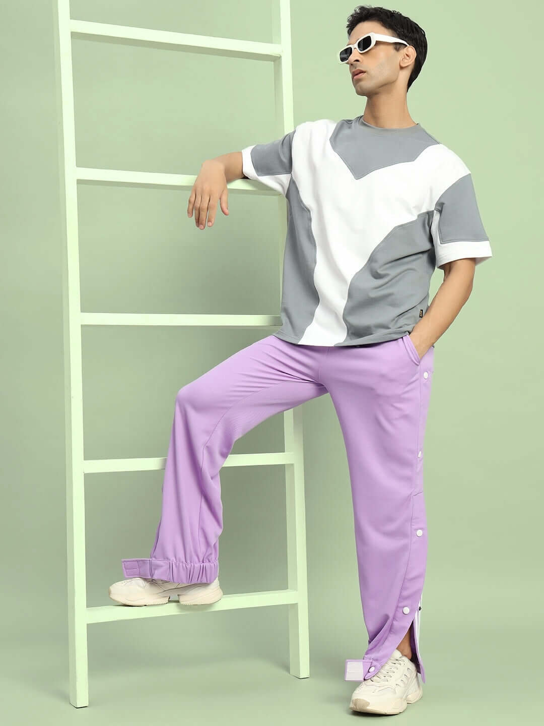 Man wearing lavender hip hop joggers and sunglasses, leaning on a mint green ladder for a stylish urban fashion look.
