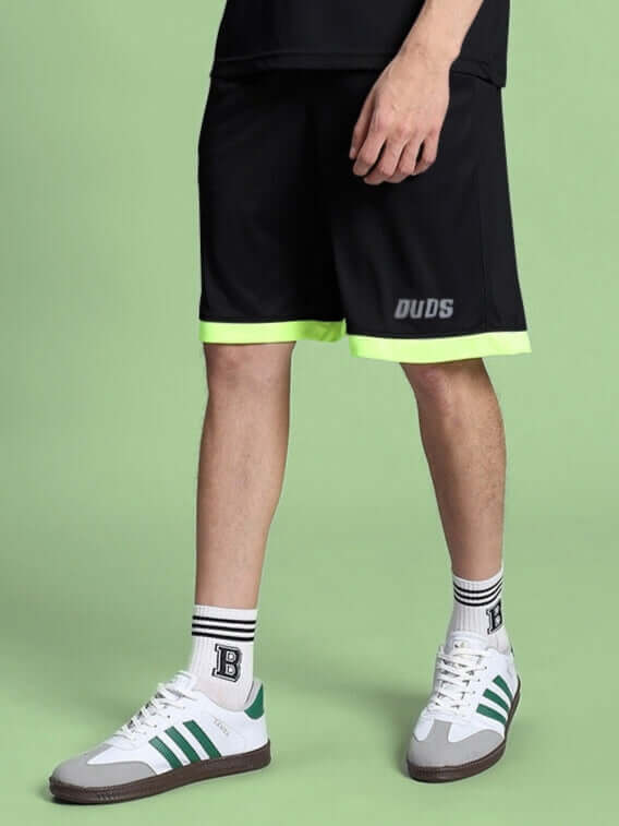 Man wearing HOOKUP REFLECTOR REGULAR FIT SHORTS BLACK with reflective details, styled with sporty socks and sneakers, on a green background.