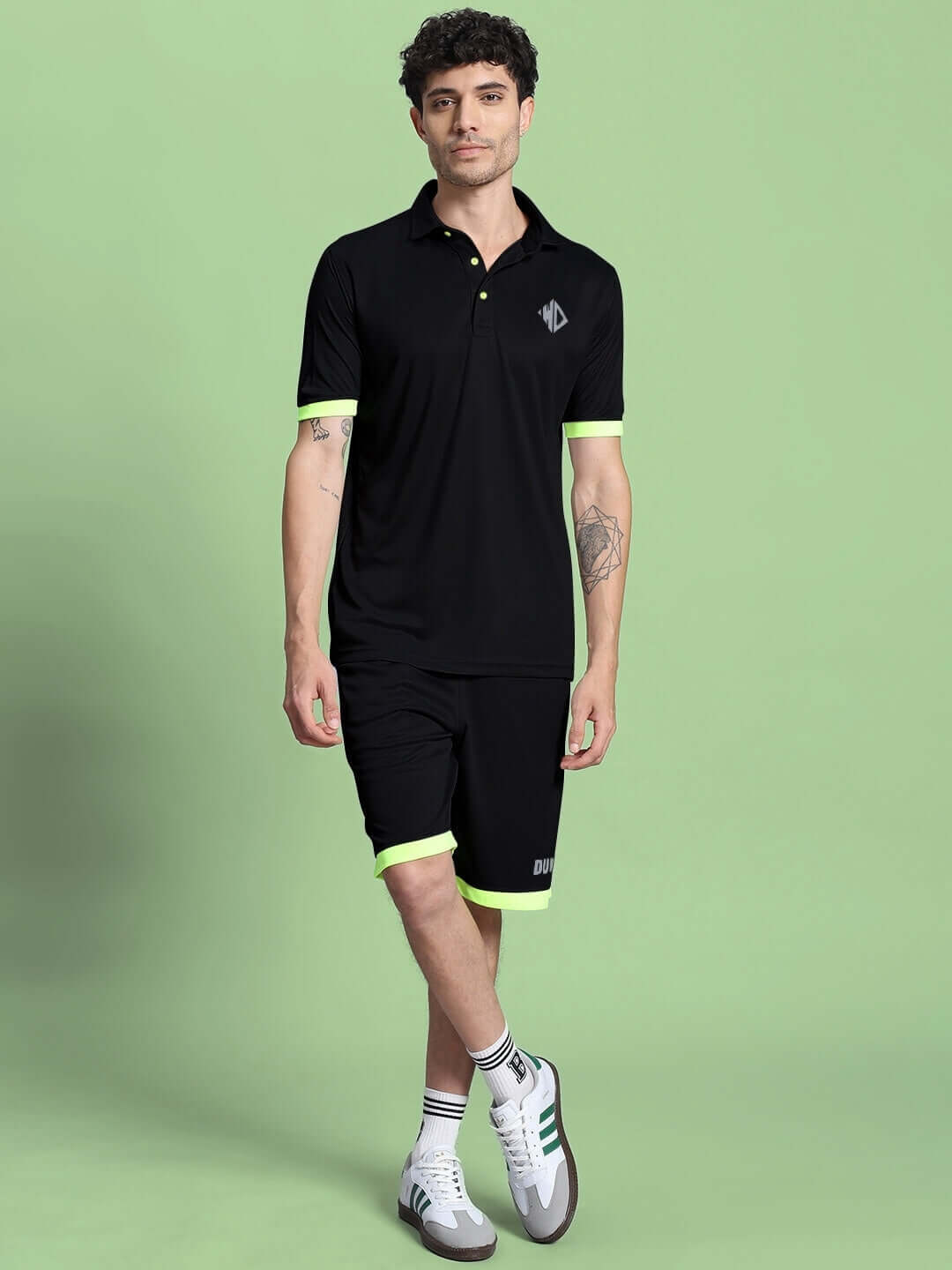 Man wearing HOOKUP REFLECTOR REGULAR FIT SHORTS BLACK with polo, standing against green background.