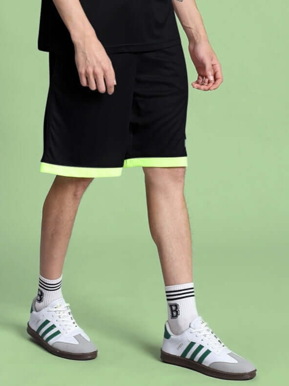 Model wearing HOOKUP REFLECTOR REGULAR FIT SHORTS BLACK with reflective details and striped sneakers on a green background.