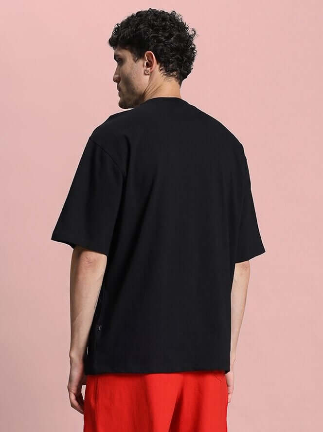 Back view of a model wearing a HOPE PRINTED OVERSIZED T-SHIRT BLACK with red pants, showcasing the oversized fit.