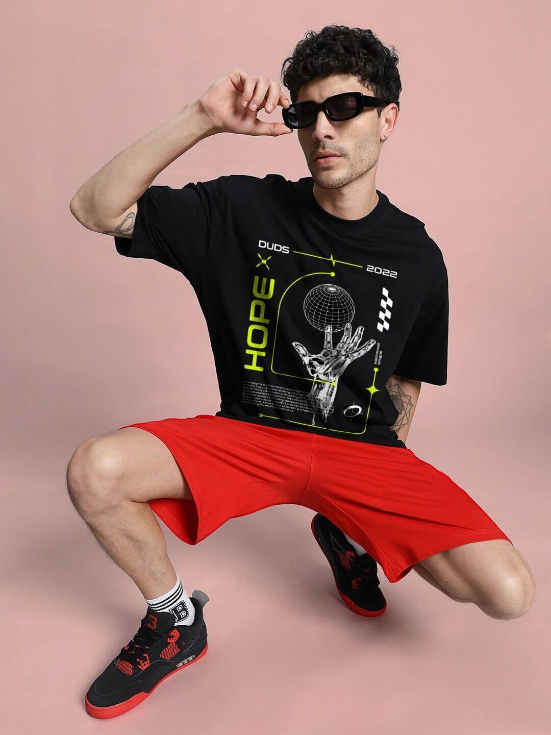 Man wearing HOPE PRINTED OVERSIZED T-SHIRT BLACK with bold graphic design, paired with red shorts and stylish sneakers.