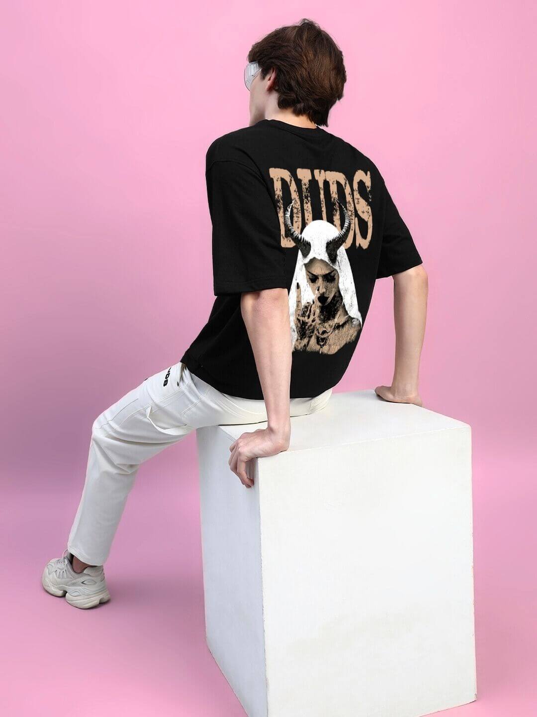 Model wearing Horned Figure Creative Print Oversized T-Shirt Black, sitting on a white cube against a pink background, showcasing the back design.