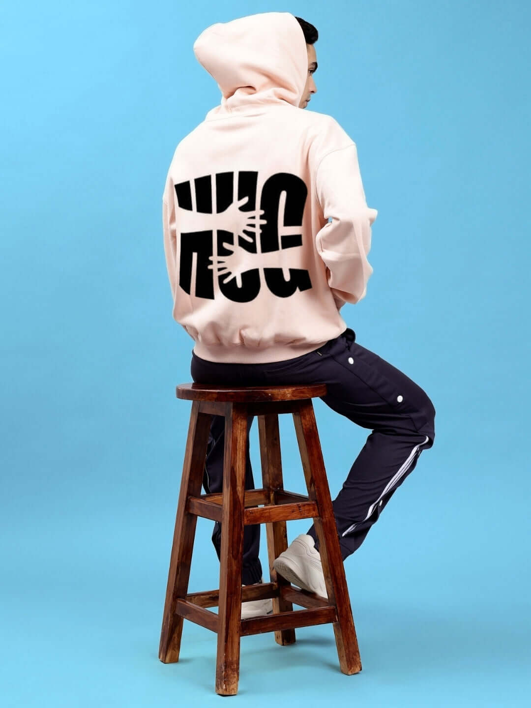 Person wearing HUG OVER-SIZED COTTON FLEECE HOODIES BEIGE sitting on a stool, showcasing back design against a blue background.