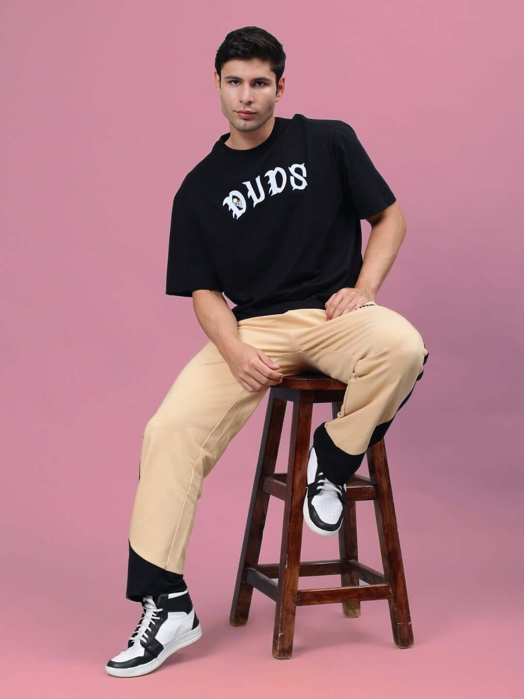 Model wearing HURTLE BAGGY JOGGERS in beige-black, paired with a black DUDS t-shirt, seated on a wooden stool against a pink background.