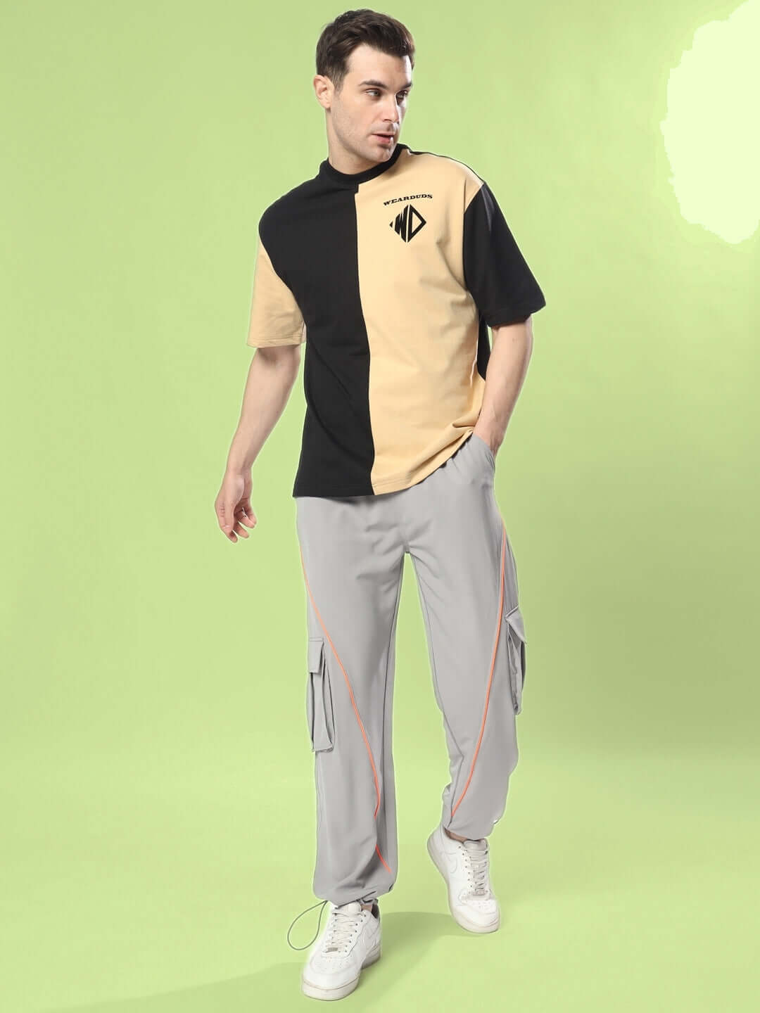 Man wearing grey cargo pants with neon orange accents and a color-blocked t-shirt on a green background.