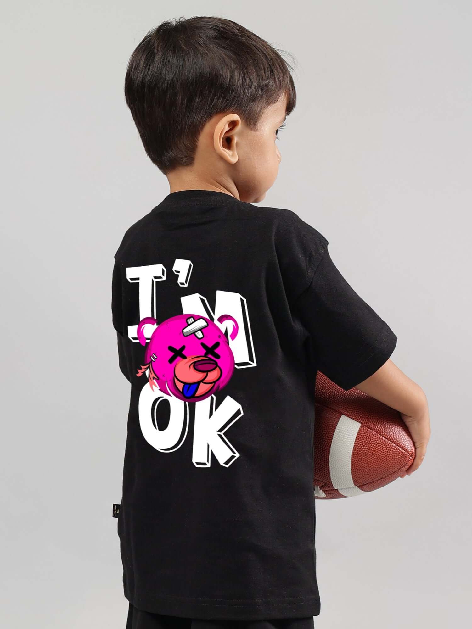 Young child wearing a black I'M OK T-shirt featuring a playful cartoon design, holding a football, perfect for boys and girls.
