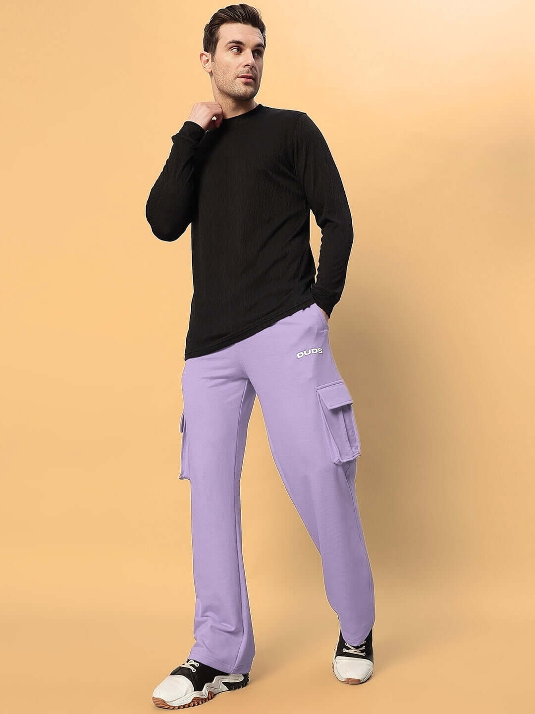 Man wearing JESS 5 Pocket Relaxed Fit Joggers in Lavender from DUDS, standing against a tan background.