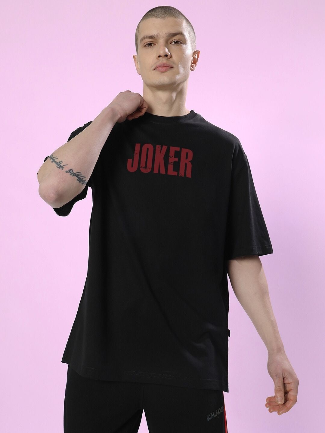 Model wearing a JOKER PRINTED OVERSIZED T-SHIRT BLACK with red lettering on a pink background.