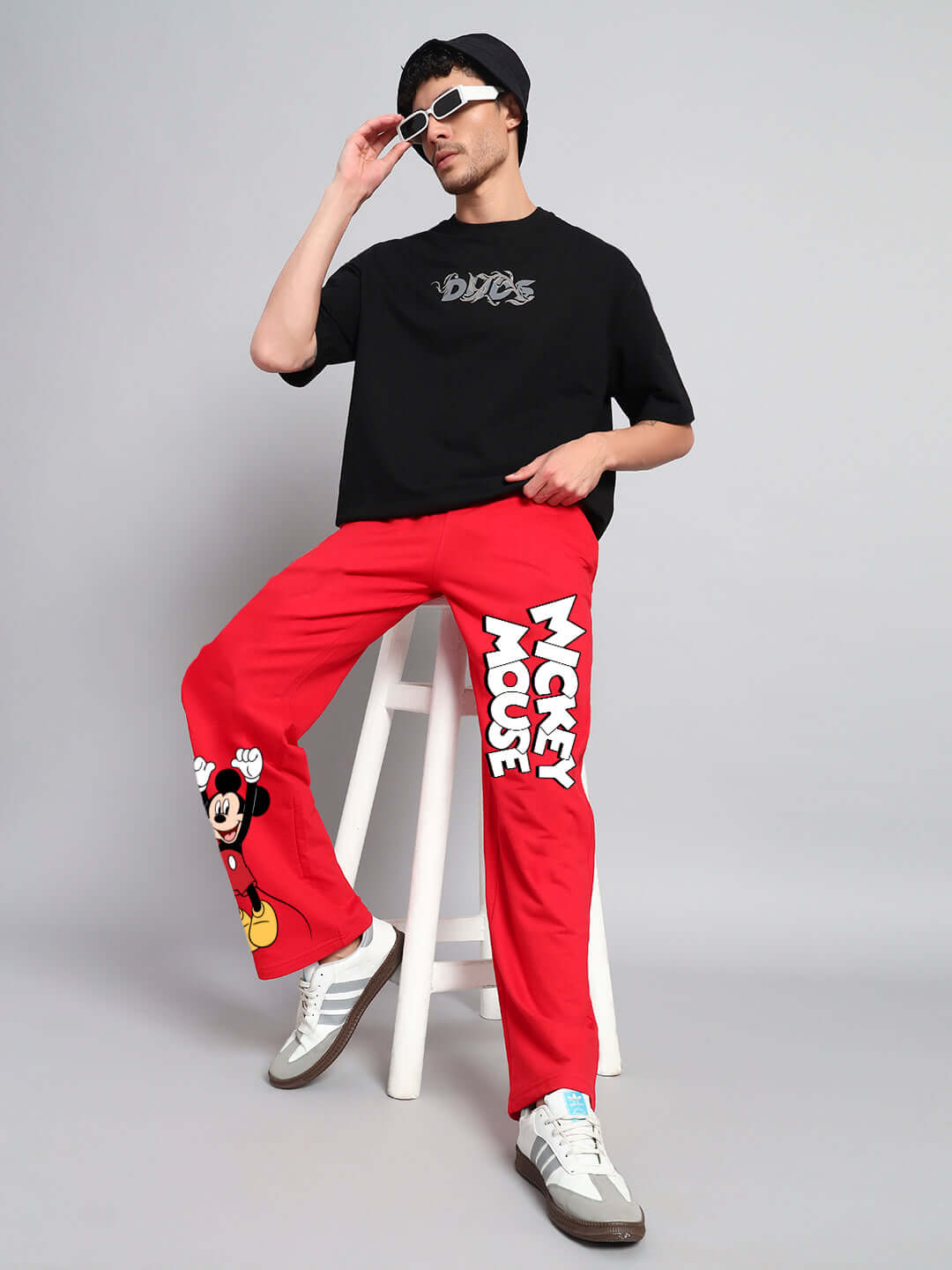 Model wearing JOY FEEL Mickey Mouse red jogger with relaxed fit, sitting on stool, sporting sunglasses, and black DUDS t-shirt.