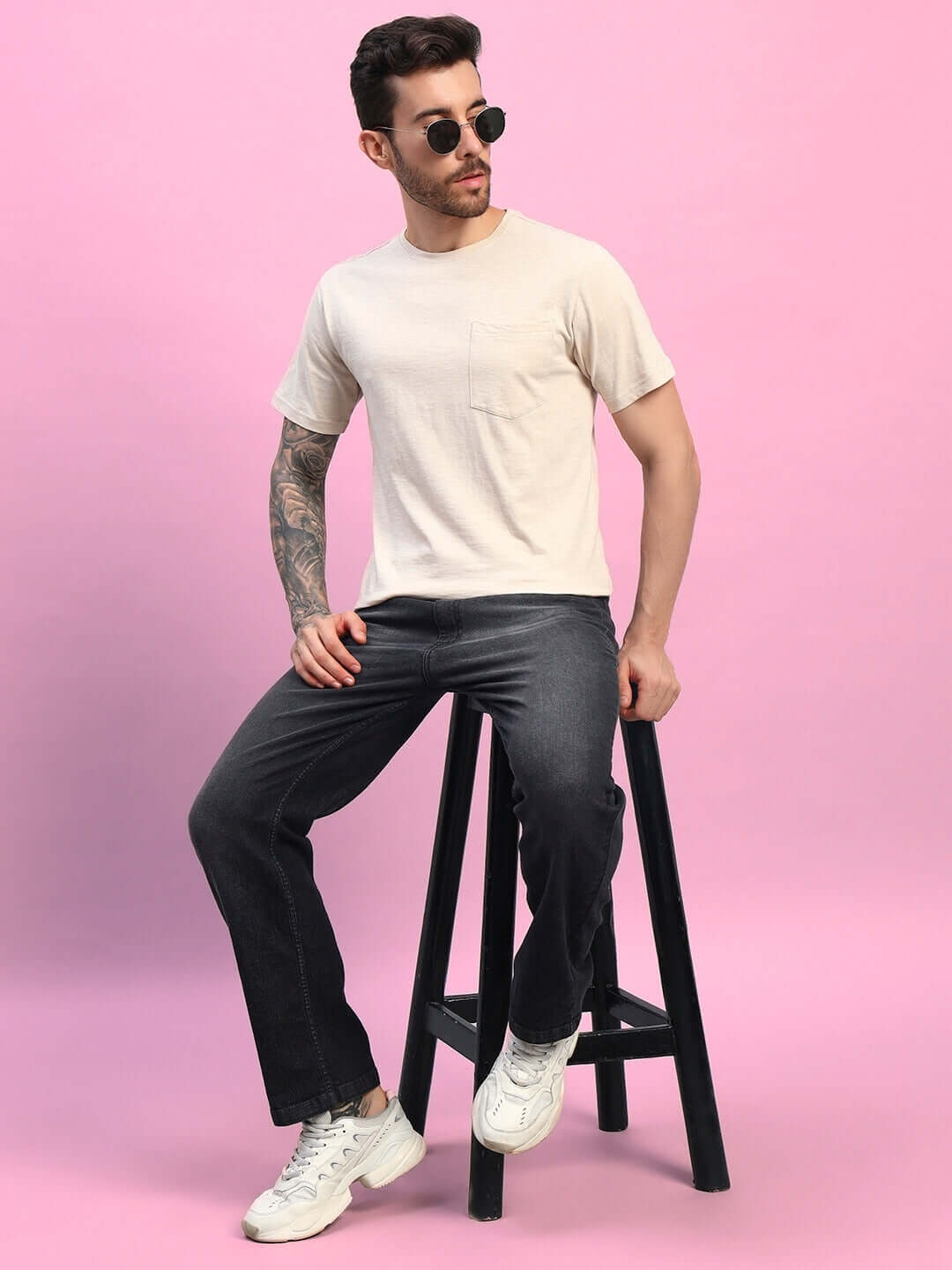 Man wearing JOY Regular Fit T-Shirt in beige, sitting on a stool, paired with black jeans and white sneakers against a pink background.